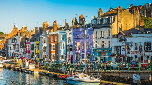 Une promenade en bord de mer bordée de bâtiments aux couleurs vives, un quai où sont amarrés des bateaux et une enseigne indiquant « Fish & Chips traditionnel ».