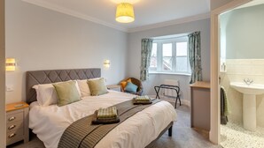 Une chambre à coucher comprenant un lit, des tables de chevet, une fenêtre avec des rideaux, un radiateur et une salle de bain avec un lavabo.