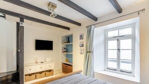 A bedroom with a bed, a TV, a window with curtains, a shelf with books, and a small table. - 1 Bed in Looe (oc-l30002) (Looe)