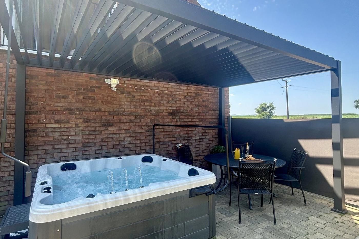 A hot tub under a covered patio with a brick wall backdrop, a table with chairs, and a clear view of the sky.