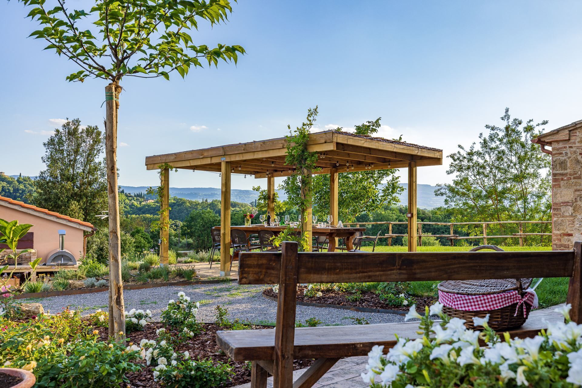 A wooden pergola with a table and chairs, surrounded by a garden with flowers and a view of the countryside.