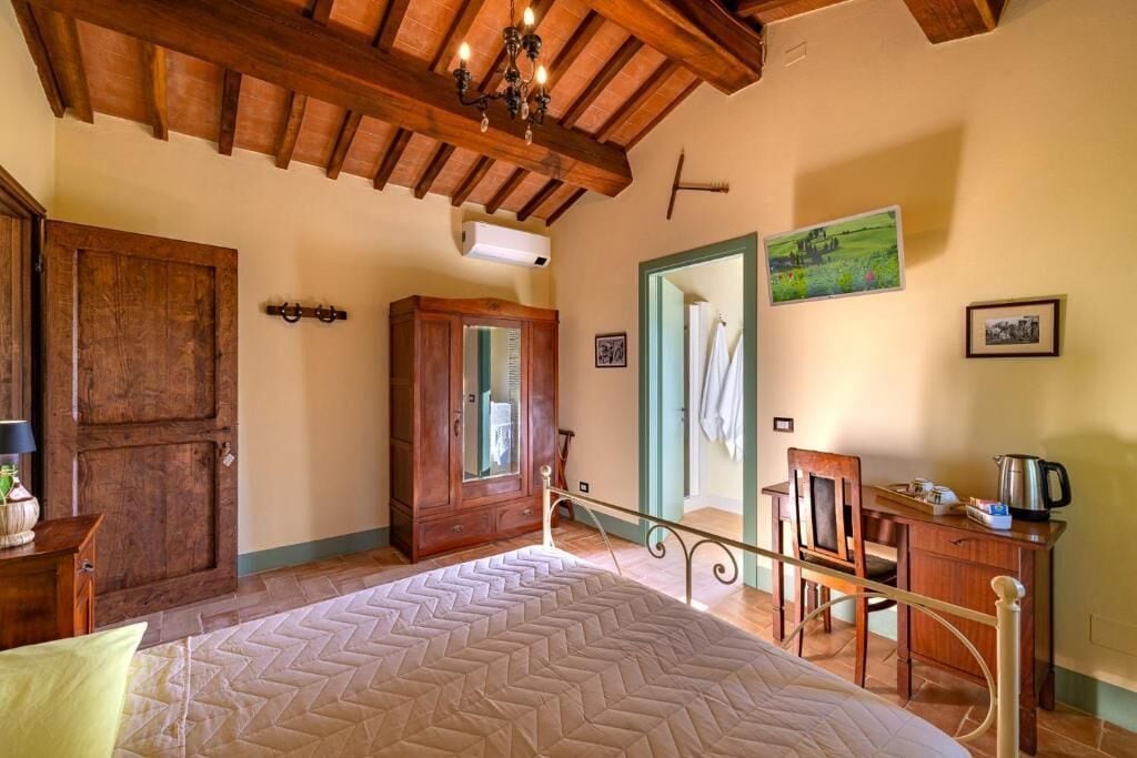 Une chambre avec un plafond en bois, un lit recouvert d’une couvre-lit à motifs, une armoire en bois, un bureau avec une chaise et un tableau accroché au mur.