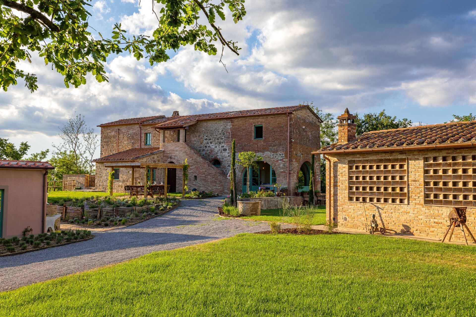 A rustic stone house with a paved driveway leading to a wooden pergola.