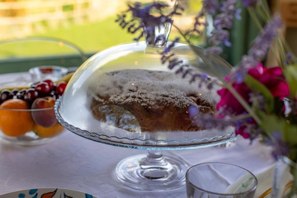Un postre sobre un expositor de vidrio, cubierto por una campana de cristal, junto a un vaso de agua y un bol de fruta.