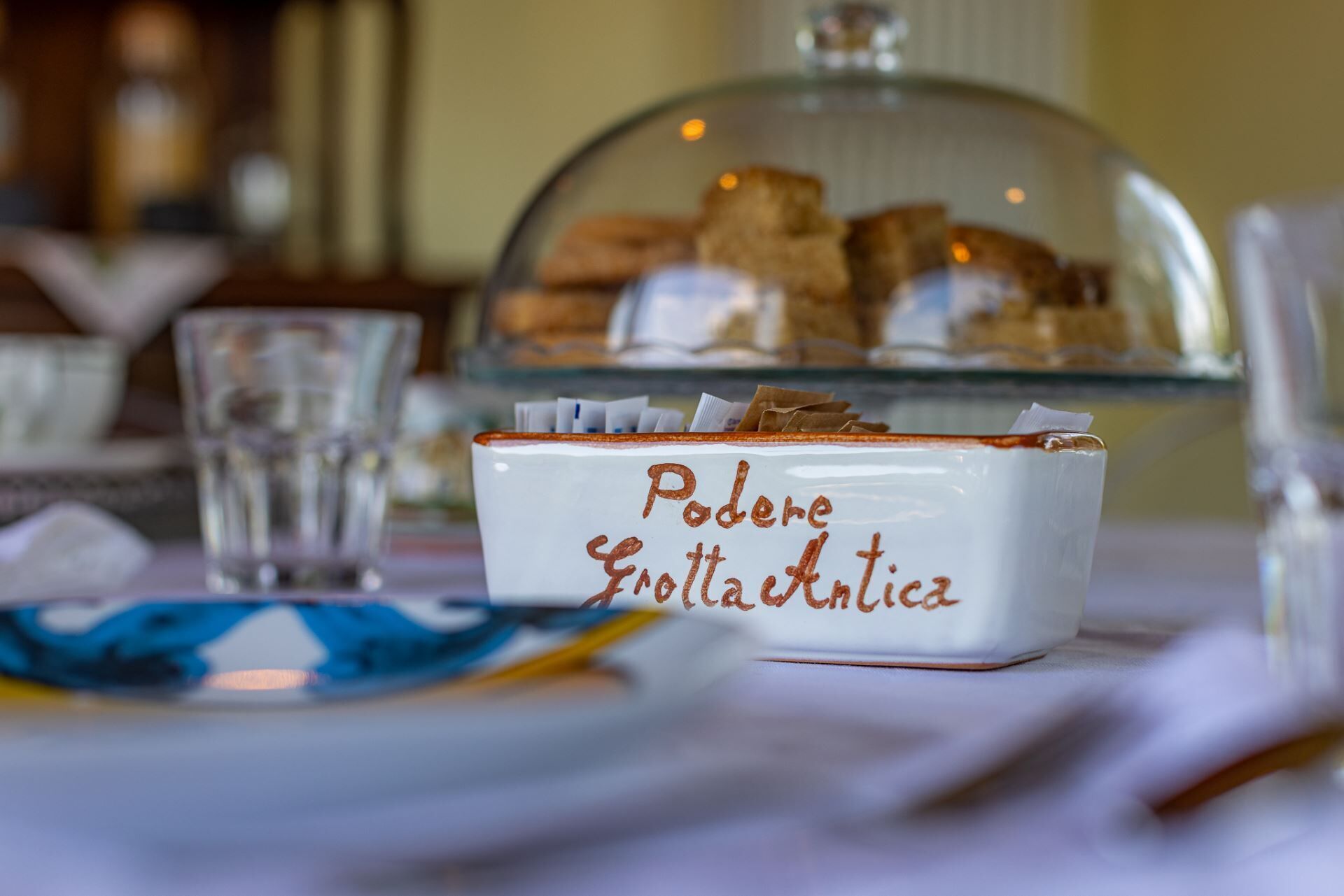 A white ceramic dish with 'Pudere Frottata Antica' written on it, placed on a table with a glass of water and a blue and white patterned plate.