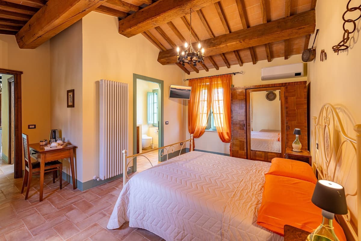 Un dormitorio con cama, cabecera de madera, mesita de noche, silla, radiador, televisor y ventana con cortinas.