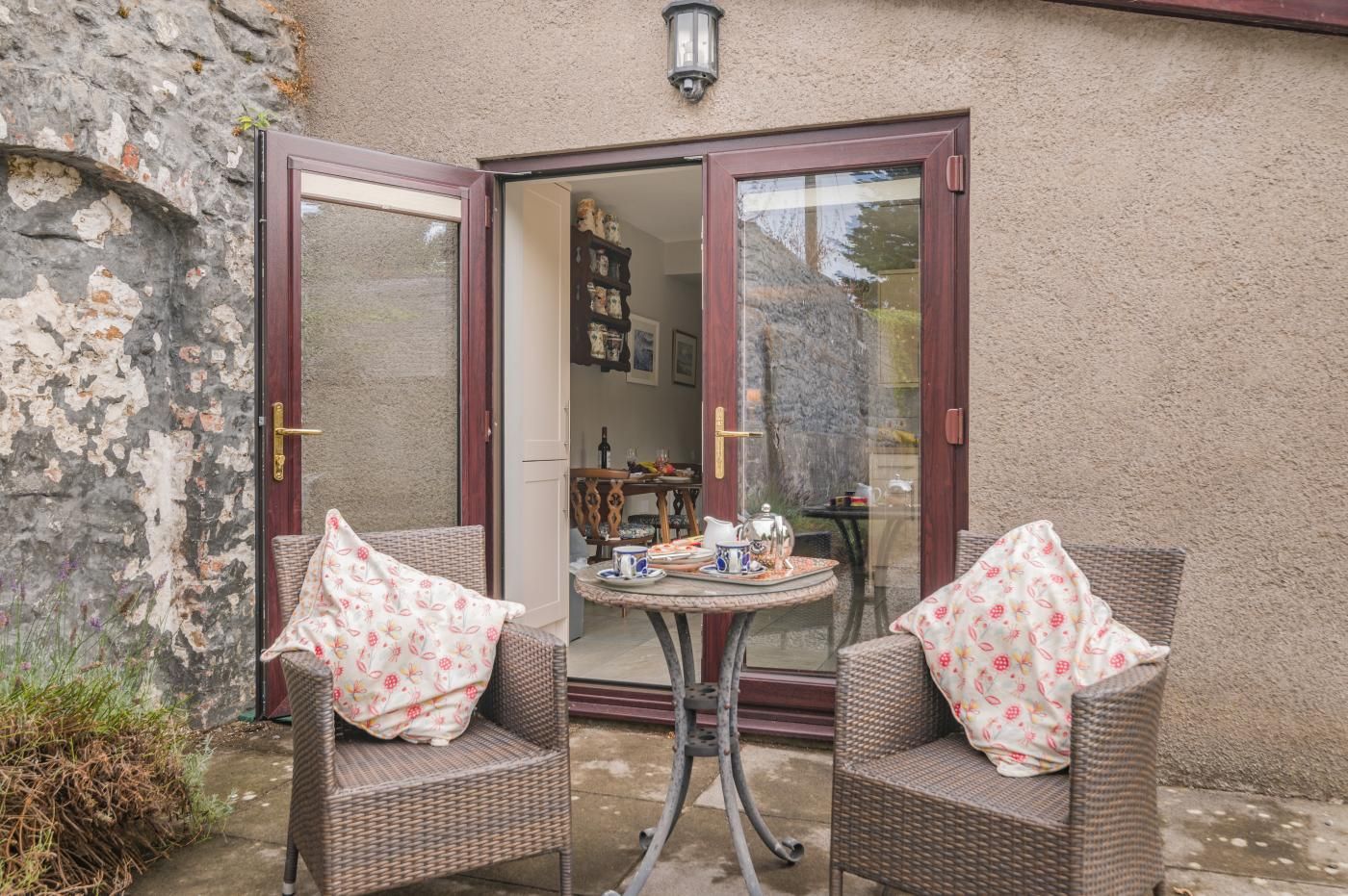 Eine Terrasse mit Korbstühlen und einem Tisch, eine Tür, die zu einem Raum führt, und eine Steinmauer.