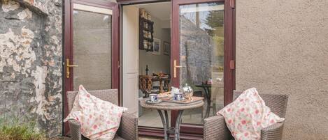 Eine Terrasse mit Korbstühlen und einem Tisch, eine Tür, die zu einem Raum führt, und eine Steinmauer.