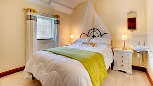 A bedroom with a bed, bedside tables, a nightstand, a window with curtains, a bedside lamp, a mirror, and a sink. - 3 Bed in Abersoch (oc-25959) (Llangian)
