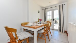 A dining area with a wooden table and chairs, a clock on the wall, and a view through the glass door. - 3 Bed in Abersoch (oc-25959) (Llangian)