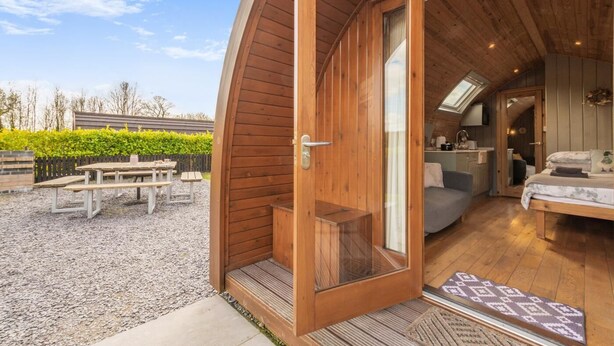 Eine Holzhütte mit großer offener Tür, die zu einem Schlafzimmer mit Bett und einer kleinen Küchenzeile führt.