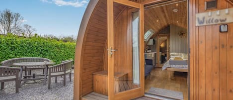 Une cabane en bois à l’extérieur incurvé, avec une porte vitrée donnant sur une chambre à coucher équipée d’un lit et d’un bureau, et un petit espace extérieur aménagé avec une table et des chaises.