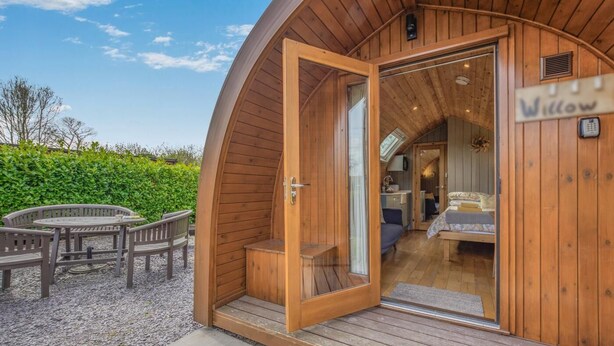 Eine Holzhütte mit gewölbter Außenwand, eine Glastür, die zu einem Schlafzimmer mit Bett und Schreibtisch führt, und eine kleine Sitzgelegenheit im Freien mit Tisch und Stühlen.
