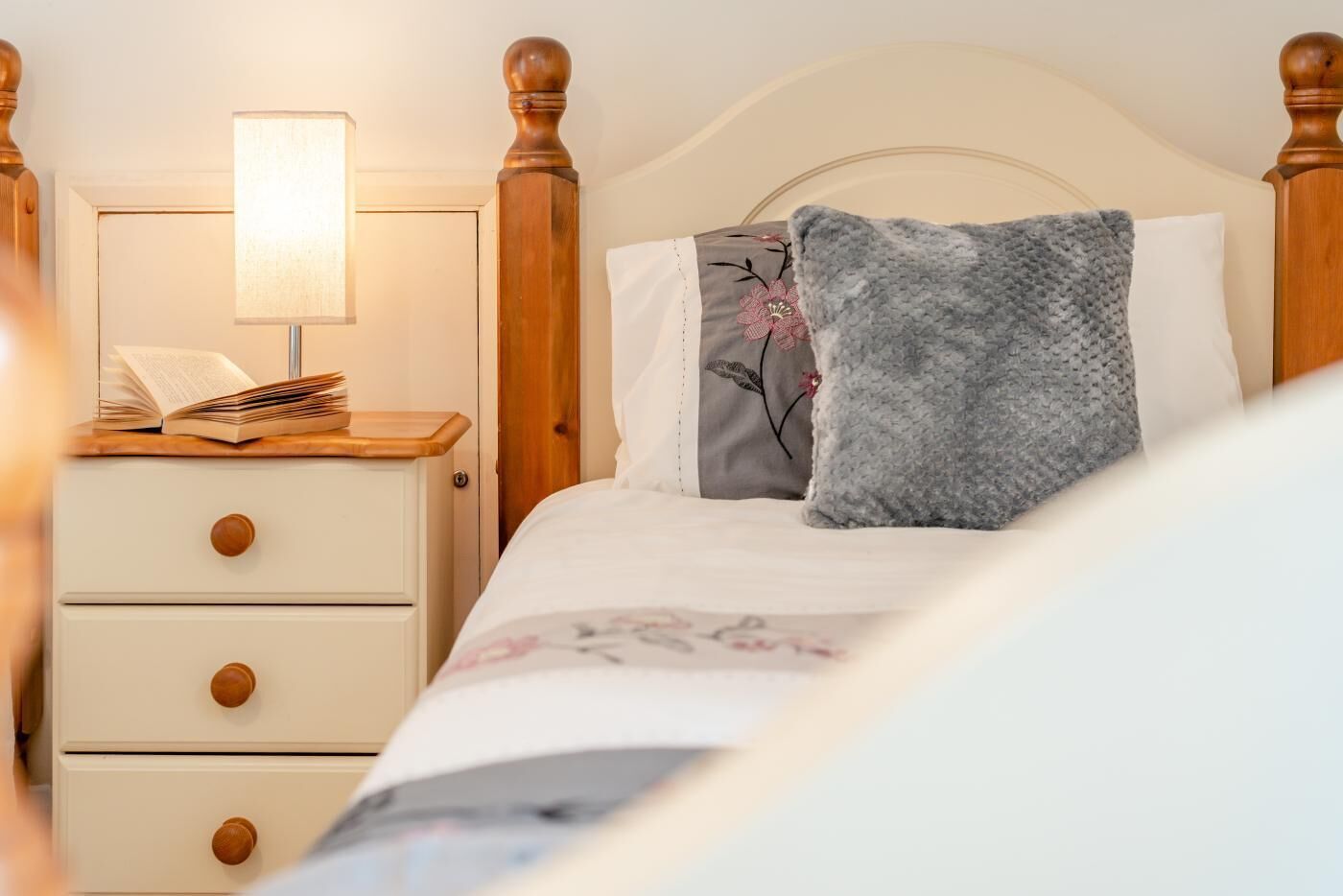 A neatly made bed with a floral bedspread, a bedside table with a lamp, and a nightstand with a book and a lamp.