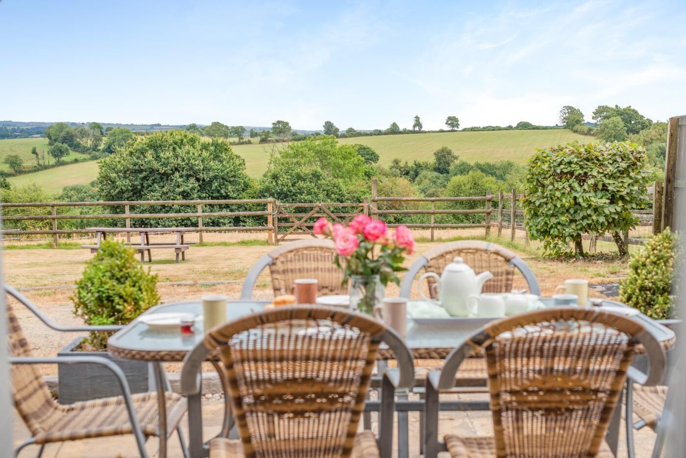 An outdoor dining area with wicker chairs and a table set for two, overlooking a rural landscape with fields and trees.