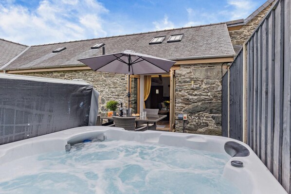 A hot tub with a table and chairs, a covered grill, and a stone building with a sloped roof.