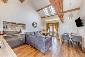 Eine moderne Küche mit grauem Sofa, Holzbalken, einem Essbereich mit Stühlen und einem an der Wand montierten Fernseher.