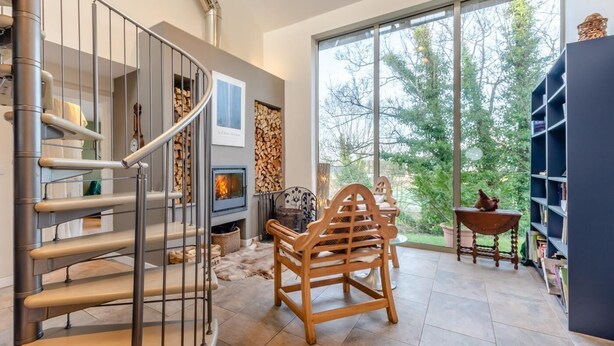 Ein modernes Interieur mit Wendeltreppe, Kamin, großem Fenster und Blick auf Bäume sowie Bücherregal.