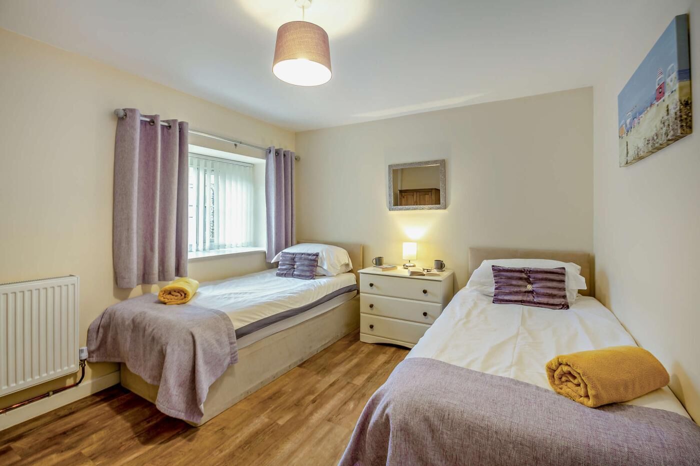 A bedroom with two beds, a window with curtains, a bedside table with a lamp, and a radiator.