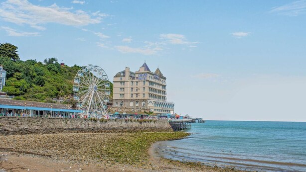 Un hotel frente al mar con una noria grande, un muelle que se adentra en el océano y una playa de rocas.