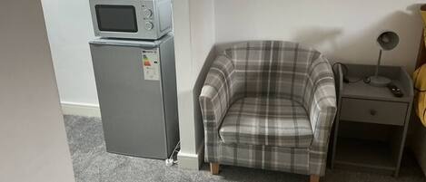 A small room with a grey refrigerator, a plaid armchair, a bedside table with a lamp, and a white wall.