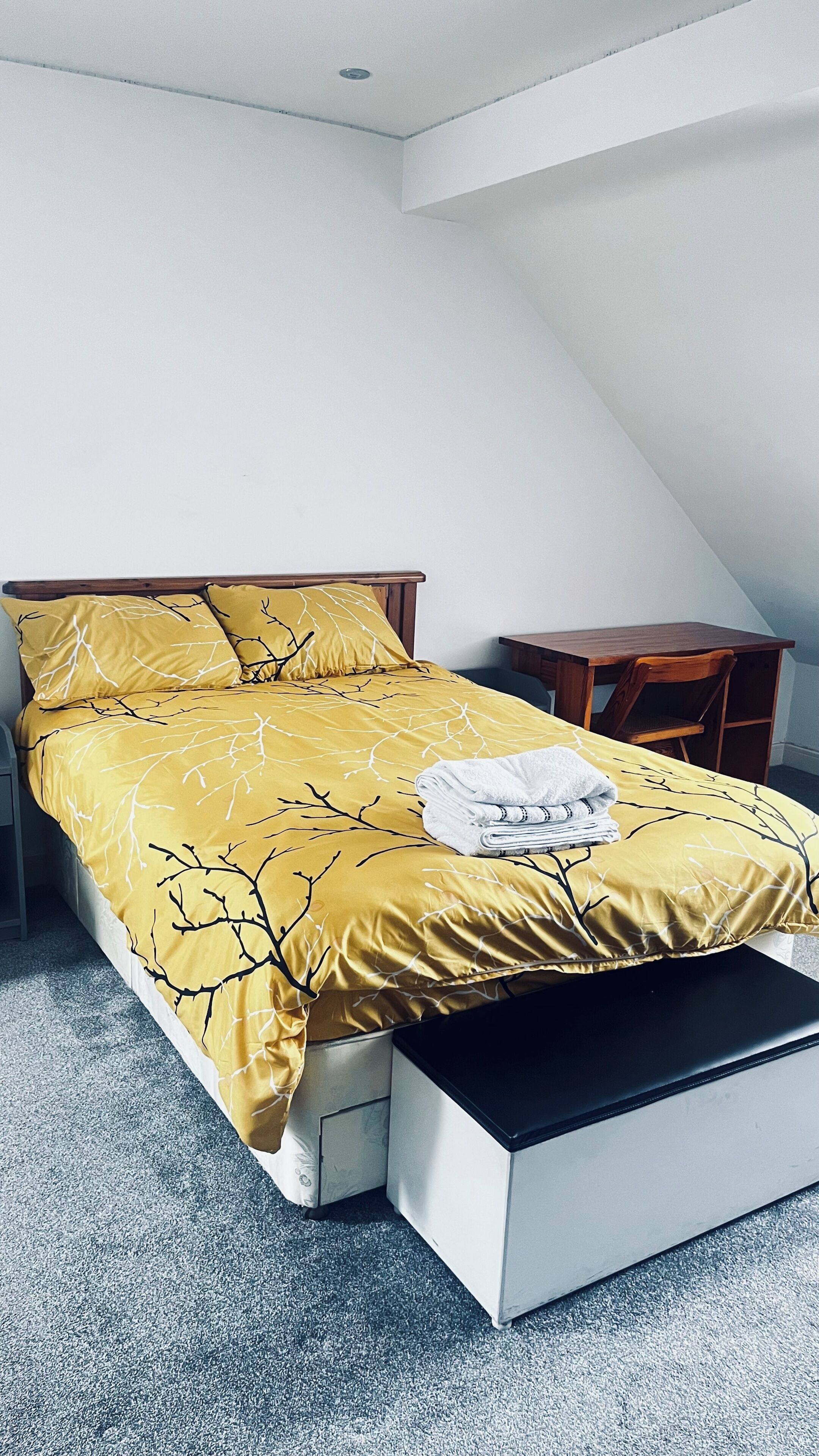 A bed with a yellow bedspread, a wooden headboard, a small wooden nightstand, and a white drawer unit.
