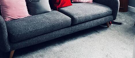 A grey sofa with multiple cushions, a wooden cabinet, and a hanging light fixture in a room with a grey carpet.