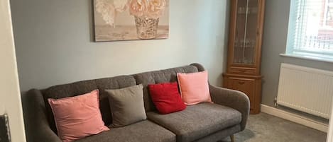 A grey sofa with pink and beige cushions, a wooden cabinet, and a painting of flowers on the wall.
