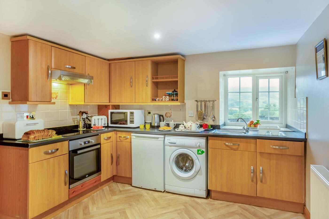 A modern kitchen with wooden cabinets, a built-in oven, a microwave, a washing machine, and a window with a view.