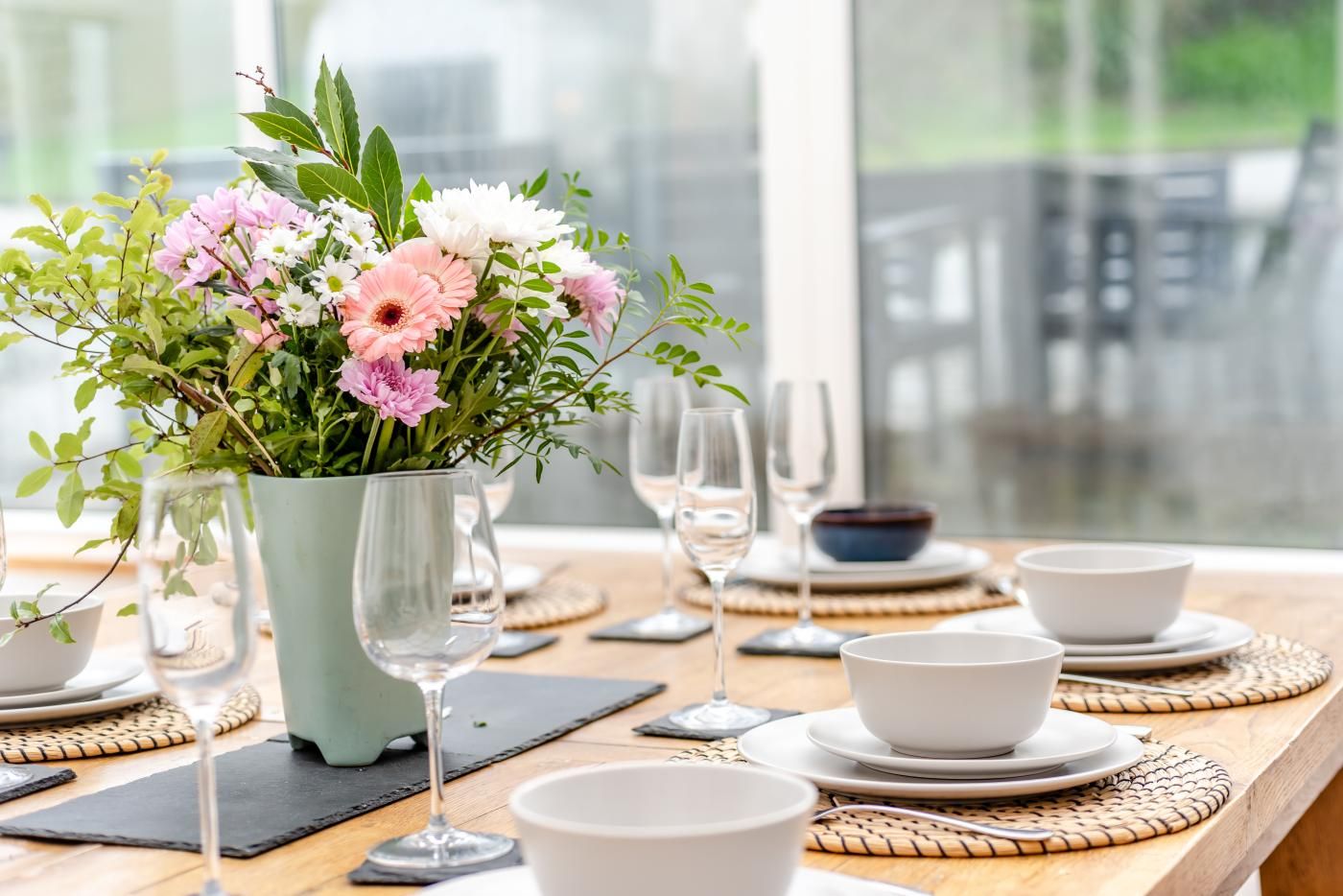 Una mesa de comedor dispuesta con cuencos blancos, copas de vino y un florero con flores rosa y blancas.