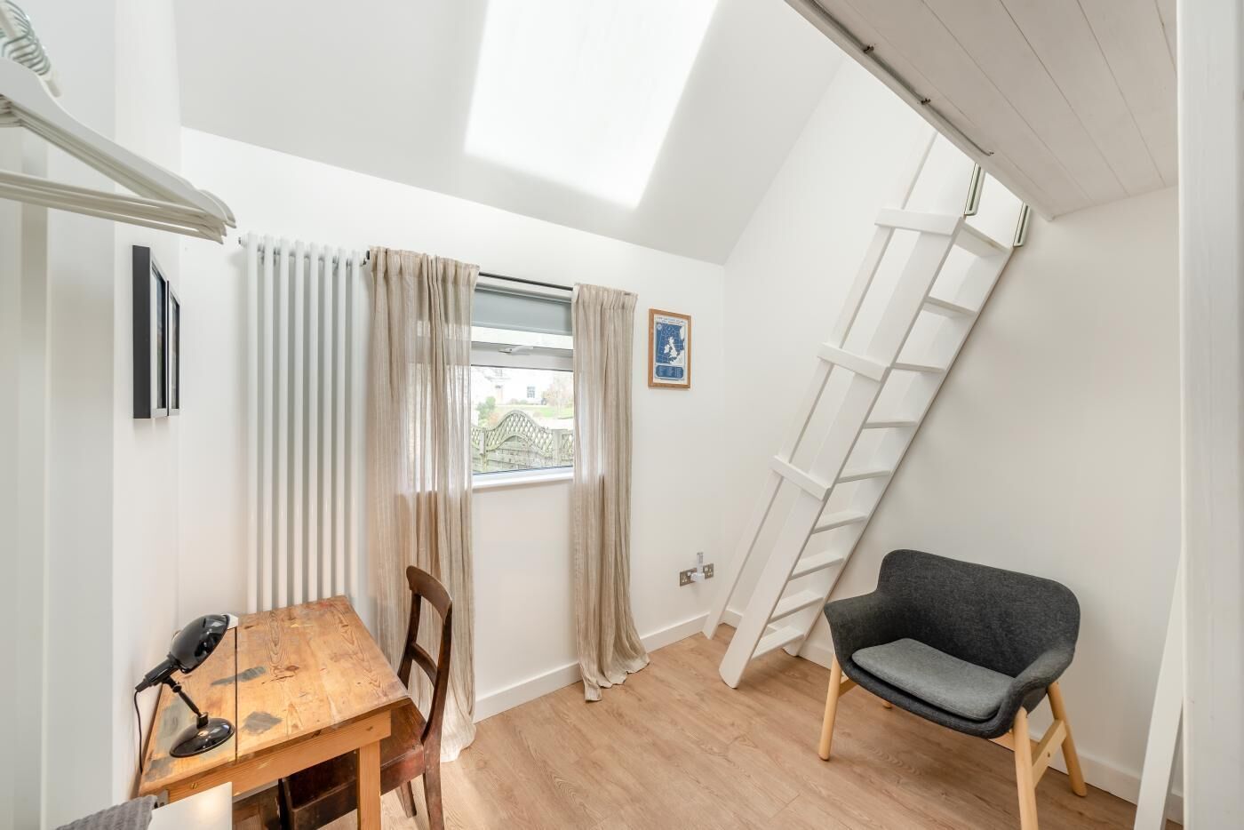 Una habitación con una mesa de madera, una silla, una lámpara de escritorio, una escalera y una ventana con cortinas.