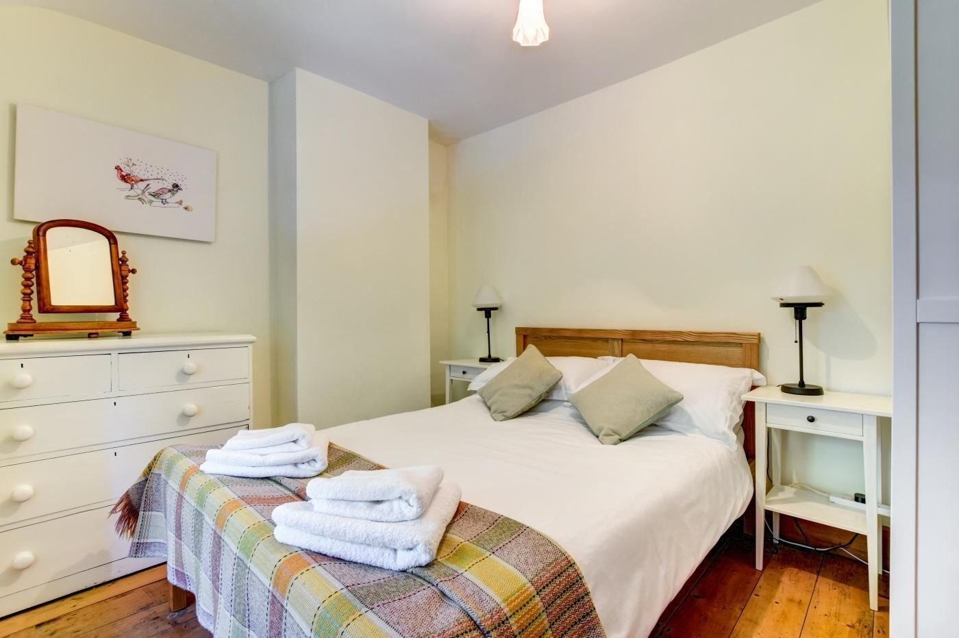 A neatly made bed with pillows, a bedside table with a lamp, a dresser with a mirror, and a framed picture on the wall.