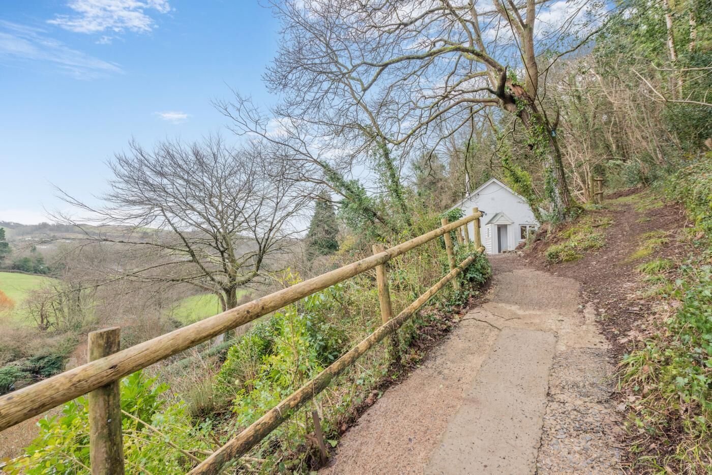 A rural path leading to a small white building with a door and windows, surrounded by trees and greenery.