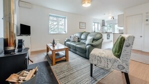 A modern living room with a sofa, a coffee table, a fireplace, and a kitchen in the background. - 2 Bed in Berrynarbor (oc-l34468) (Berrynarbor)