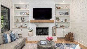 A living room with a flat-screen TV, fireplace, white shelves with decorative items, a sofa, and a coffee table. - Stay @ Isle of Coladas (Isle of Palms)