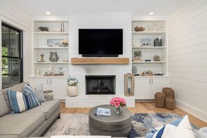 A living room with a flat-screen TV, fireplace, white shelves with decorative items, a sofa, and a coffee table. - Stay @ Isle of Coladas (Isle of Palms)