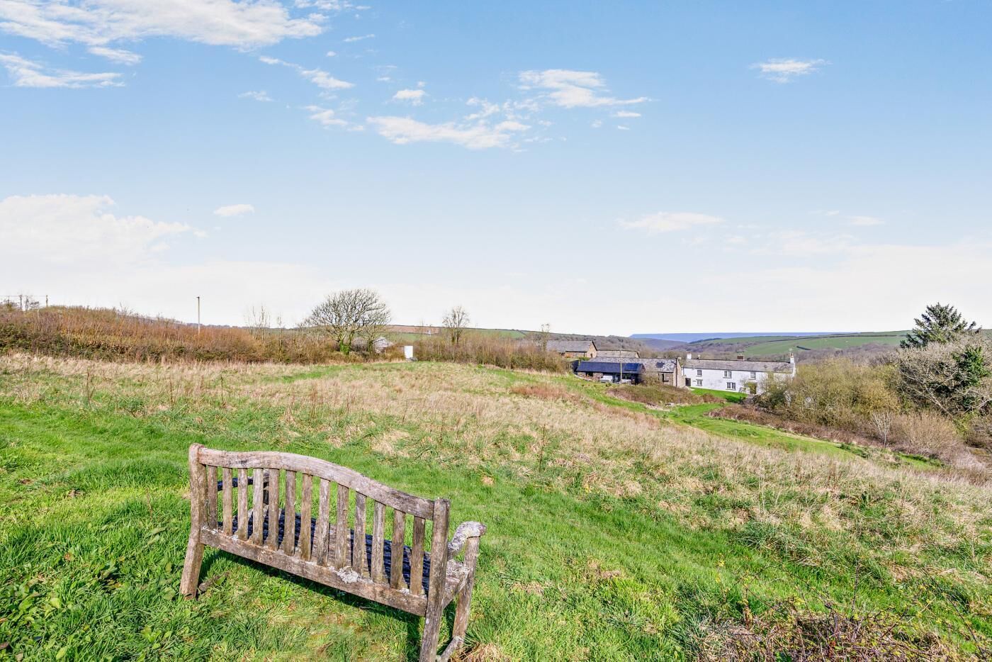 A rural landscape with a wooden bench, a white building with a blue roof, and a clear sky.