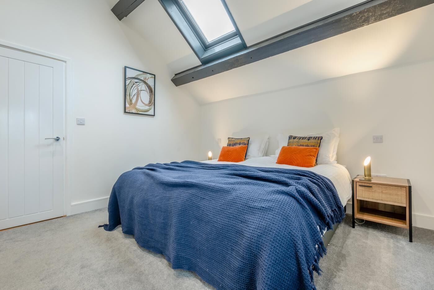 A bedroom with a skylight, a bed with a blue blanket, two orange pillows, and a bedside table with a lamp.