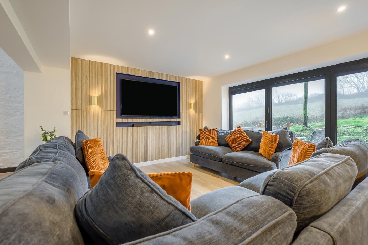 Ein modernes Wohnzimmer mit einem großen Flachbildfernseher an einer Holzwand, einem grauen Ecksofa mit orangefarbenen Kissen und einem großen Fenster mit Blick ins Freie.