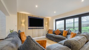 Ein modernes Wohnzimmer mit einem großen Flachbildfernseher an einer Holzwand, einem grauen Ecksofa mit orangefarbenen Kissen und einem großen Fenster mit Blick ins Freie.