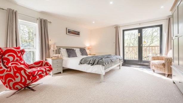 Une chambre à coucher avec un lit, des tables de chevet, un fauteuil à motifs rouges, un fauteuil à rayures et une vue à travers une porte vitrée.