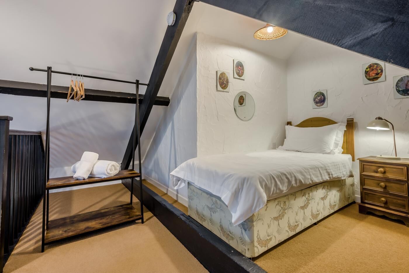 A bedroom with a bed, a wooden nightstand, a lamp, a shelf with towels, and a wall with framed pictures.