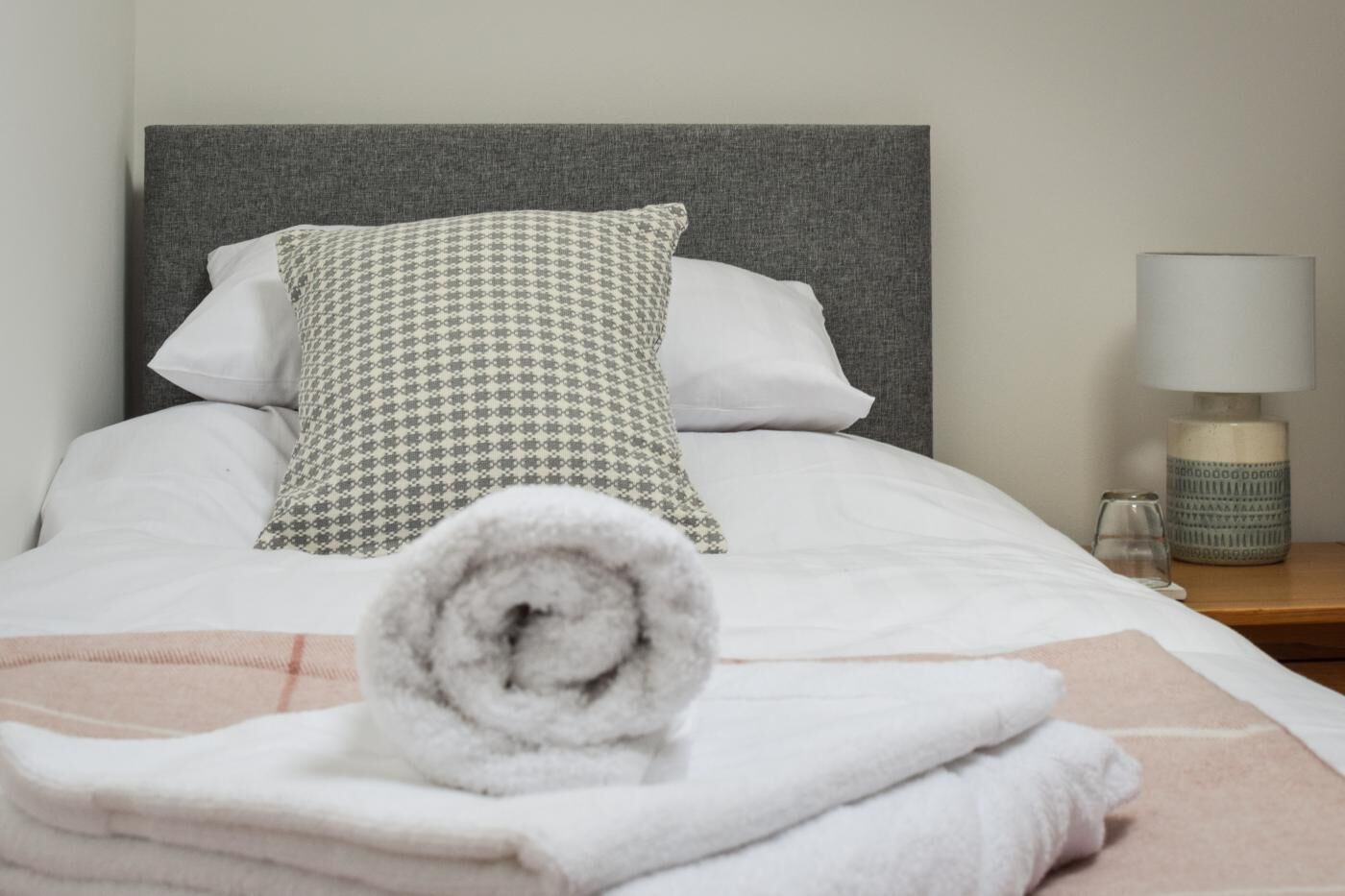 Uma cama bem arrumada com cabeceira cinza, roupa de cama branca e almofada estampada. Uma toalha enrolada está sobre a cama, e um criado-mudo com abajur e um copo são visíveis.