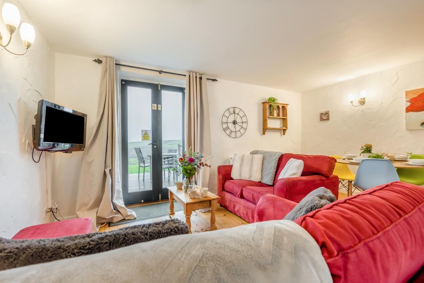 Un salon avec un canapé rouge, un téléviseur fixé au mur, une table d’appoint en bois et un coin repas avec une table dressée pour deux personnes.