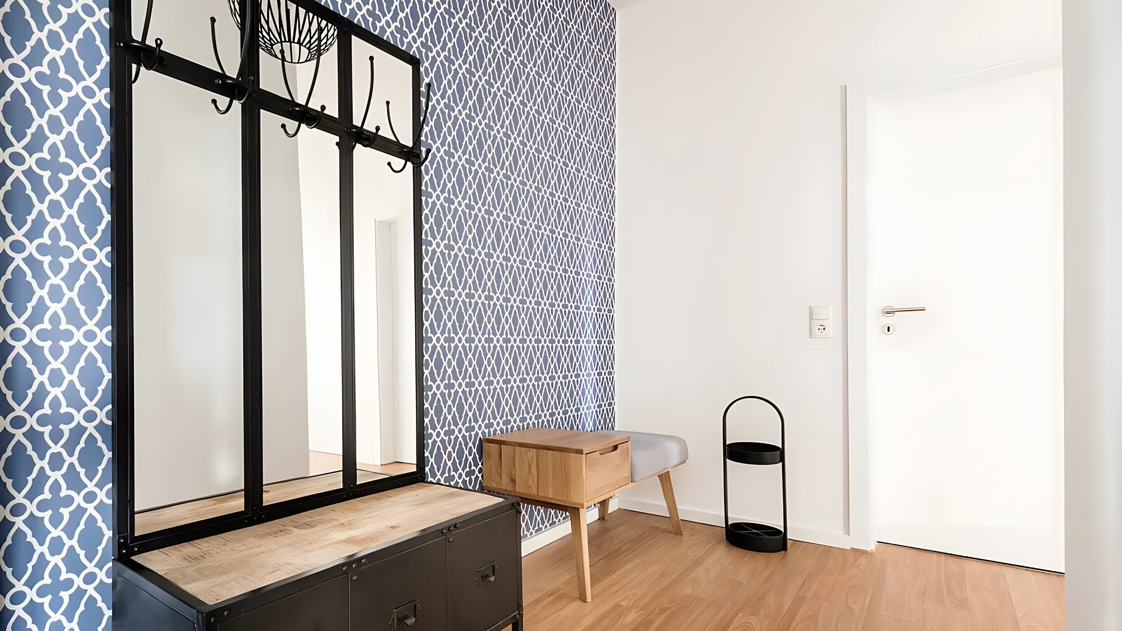 Une pièce comprenant une commode en bois, un miroir avec un cadre noir, un mur à motifs, une porte et un petit présentoir noir.