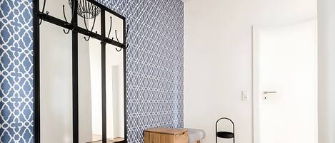 Une pièce comprenant une commode en bois, un miroir avec un cadre noir, un mur à motifs, une porte et un petit présentoir noir.