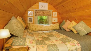 A cozy, wooden interior with a bed, pillows, a window, and framed pictures on the wall. - Daisy Glamping Pod (Newbury)