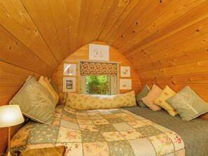 A cozy, wooden interior with a bed, pillows, a window, and framed pictures on the wall. - Daisy Glamping Pod (Newbury)