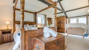 A four-poster bed with a wooden headboard, a wooden chest of drawers, a small bedside table with a lamp, a sofa, and a large window with curtains. - 1 Bed in St. Gennys (oc-v34634) (Crackington Haven)