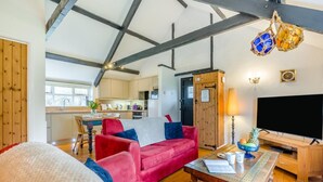 A living room with a red sofa, a wooden coffee table, a TV, and a kitchen in the background. - 1 Bed in St. Gennys (oc-v34634) (Crackington Haven)